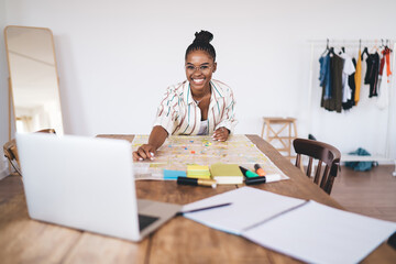 Cheerful African American woman enthusiastically planning trip using paper map, smiling near laptop...