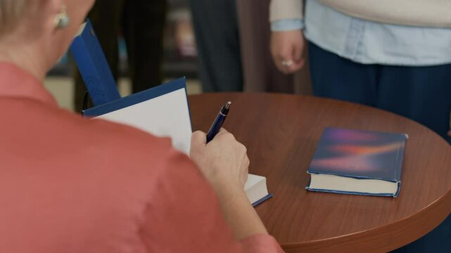 Ethnically diverse bookstore attendees hold hardcover books as they waiting in line for signature of mature Caucasian female author, sitting at round table with pen