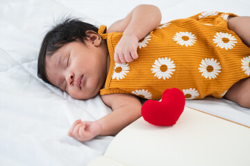 Mixed race, Asian and African, newborn one month baby girl sleeping on bed lying on white sheets with a small red heart pillow and book. Close up. Side view