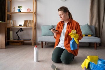 Woman housewife sitting with cleaning supplies on floor reading news and social media in her smartphone