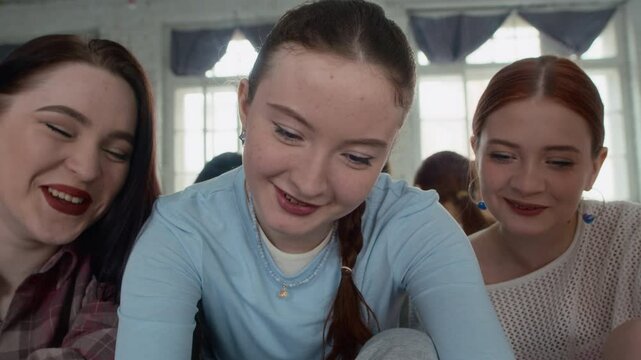 Tilt up shot of Caucasian female zoomers watching their dance filmed on mobile phone sitting on floor after training in studio
