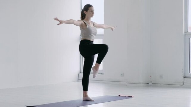 Gorgeous caucasian young female in sportswear doing warm-up before yoga In a bright studio with windows. Girl stands on one leg and rotates the other. Balance and equilibrium of the body exercise