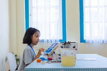 Young Asian student deeply in a painting workshop, brushing colors onto a canvas. Indoor pastel-toned classroom enhances the calm, focused mood. Surrounded by tools and paints for hands-on learning.
