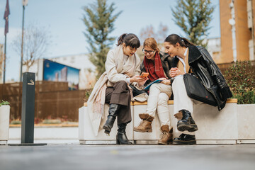 Three businesswomen are seated close together on a bench, sharing a fun moment as they look at a smart phone. The scene shows camaraderie and the joy of friendship in an urban setting.