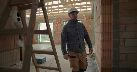 Construction worker with helmet walking through house under construction, observing progress, surrounded by exposed brick walls, wooden beams, and window frames