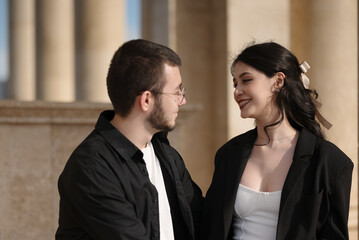 Happy couple sharing a joyful moment together outdoors near classical columns. The woman smiles warmly while looking at her partner, conveying romance and connection.