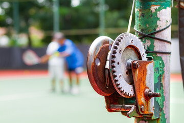 Old and rustic tennis net reel post with blurred persons taking tennis lesson on the background