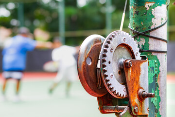 Old and rustic tennis net reel post with blurred persons taking tennis lesson on the background