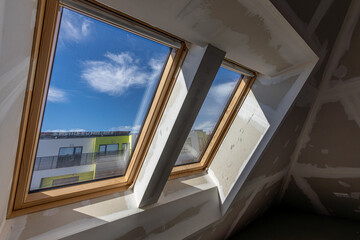 Two large skylight windows with wooden frames in an unfinished attic space under construction. Natural light enters the room, highlighting the drywall and modern architectural design...