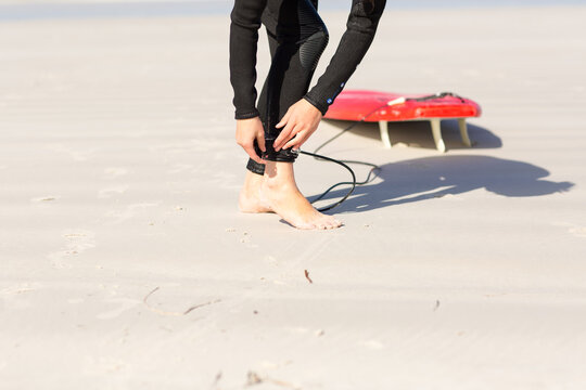 Surfer fastening ankle strap/leg rope with surfboard on the beach - Powered by Adobe