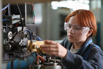 Young female engineer with red hair wearing safety glasses working on machinery in workshop, repairing industrial equipment in technical laboratory