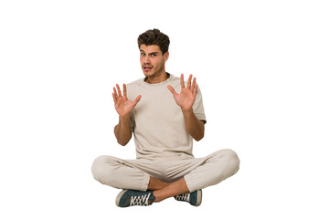 Young caucasian man sitting on the floor isolated on white background rejecting someone showing a...