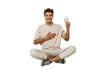 Young caucasian man sitting on the floor isolated on white background taking an oath, putting hand on chest.