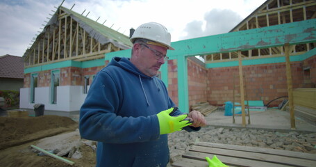 Man removing gloves and helmet at construction site, wearing glasses, with house under construction in background, preparing to finish work outdoors