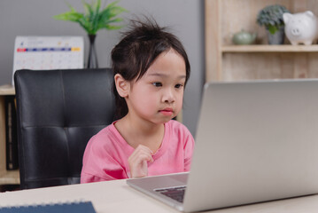 Asian child girl studying online at home using a laptop, focused on screen, representing...