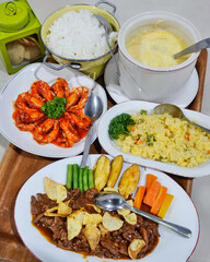 Assorted dishes including sautéed shrimp in a red sauce, white rice in a metal pot, egg soup in a large bowl, and a vegetable salad with potatoes, carrots, and broccoli