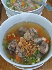 A bowl of clear soup contains pieces of meat and bone, likely oxtail, with sliced green onions and fried shallots on top