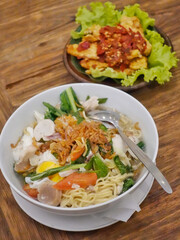 A white bowl containing a noodle soup dish is topped with vegetables, slices of meat, a boiled egg, and crispy shallots