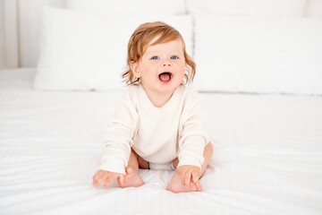 Fototapeta premium A baby with red hair and blue eyes in a white bodysuit is smiling happily on the bed at home, a healthy happy cute newborn baby boy