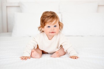 A baby with red hair and blue eyes in a white bodysuit is smiling happily on the bed at home, a...