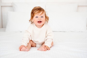 Fototapeta premium A baby with red hair and blue eyes in a white bodysuit is smiling happily on the bed at home, a healthy happy cute newborn baby boy