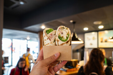 A Hand Holding a Takeaway Chicken Caesar Salad Wrap in Front of a Bustling Cafe Counter Showcases Fresh Grab and Go Dining Inside a Trendy London Eatery.