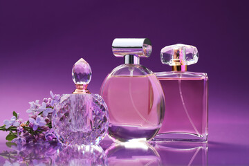 Bottles with perfumes and lilac flowers on mirror surface against color gradient background, closeup