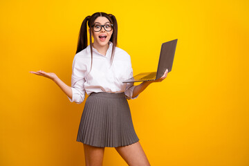 Young woman holding laptop and gesturing in front of yellow background, styled as a schoolgirl with...