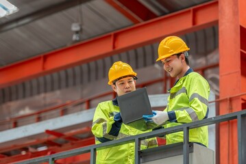 Two male construction workers wea yellow safety helmets and high visibility jackets discussing plans on a laptop at a construction site with steel framework in the background