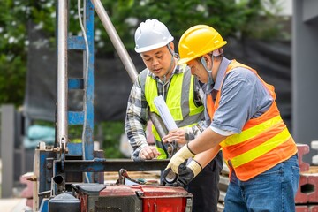 Two male construction workers wea safety helmets and reflective vests inspecting machinery on a...