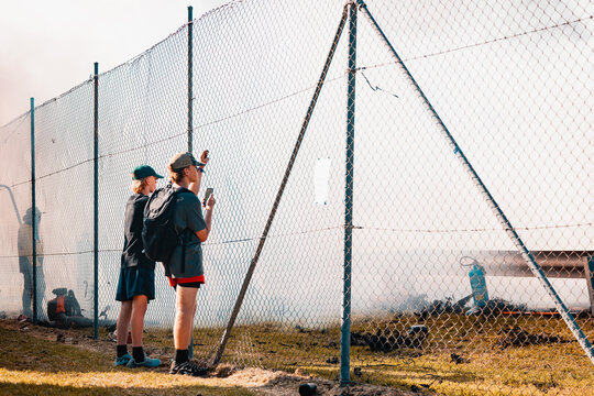 Teenagers watching vehicle burnout competition through chain mesh fence with smoke haze from burning