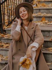 Beautiful blonde young woman in hat and coat sitting on the stairs in the autumn park.