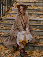 Beautiful blonde young woman in hat and coat sitting on the stairs in the autumn park.