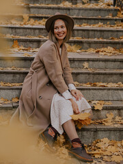 Beautiful blonde young woman in hat and coat sitting on the stairs in the autumn park.