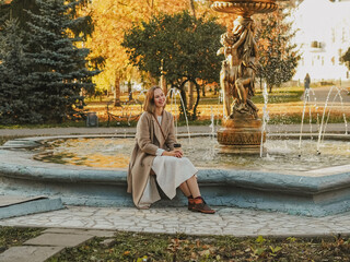 Beautiful blonde young woman in hat and coat drinking coffee and walking in autumn city