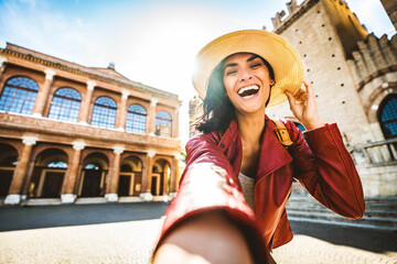 Happy young woman with hat and backpack taking selfie portrait with smart mobile phone in city street - Delightful female tourist enjoying summer vacation walking in old town
