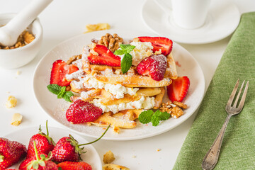 Belgian waffles with cottage cheese cream, strawberries, walnuts and mint on a white plate on a light table with a cup of coffee. Delicious, Sunday breakfast.
