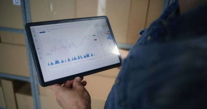 Employee Checking Real-Time Stocks and Exchange Market Charts Using Digital Tablet Computer. Man Making Investments During Working Hours in Warehouse, Combining Job with Online Trading. Close Up Shot. - Powered by Adobe