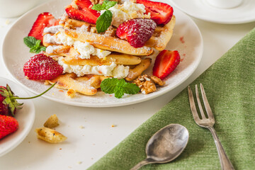 Belgian waffles with cottage cheese cream, strawberries, walnuts and mint on a white plate on a light table with a cup of coffee. Delicious, Sunday breakfast.