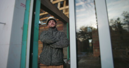 Young construction worker with arms crossed, wearing a hard hat, looking determined, standing by a glass wall with wood framework in the background on a construction site
