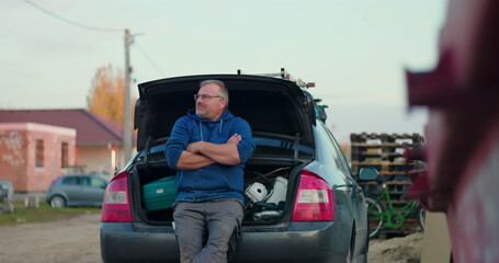Worker leaning on car trunk at construction site, arms crossed and observing surroundings, emphasizing thoughtfulness and preparation during renovation project