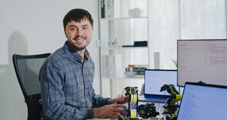 Portrait confident smiling male programmer working with robotics code AI software and looking at camera in tech office. Artificial intelligence dashboard, robotics, user interface, machine learning.