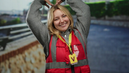 Volunteer woman with blonde hair making heart symbol on city street, wearing red vest and smiling...