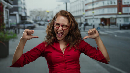 Woman in red shirt smiling and pointing at herself energetically on a city street depicting...