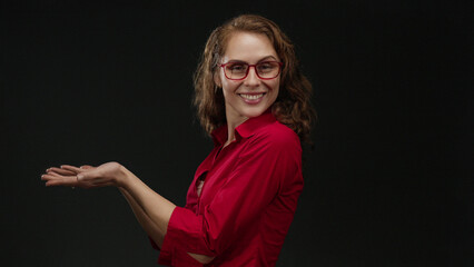 Woman smiling in a red shirt with extended arms over an isolated black background, offering a sense...