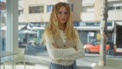 Woman standing confidently with arms crossed in a bright indoor office setting, wearing glasses and...