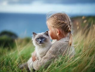 Grey ragdoll cat and girl holding him and sitting on green grass in spring landscape with sea in the background, view in natural light with shallow depth of field, realistic outdoor animal portrait