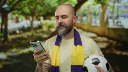 Fototapeta premium Bald man with beard holds smartphone and soccer ball, wearing yellow and purple scarf, standing in a sunny park setting.