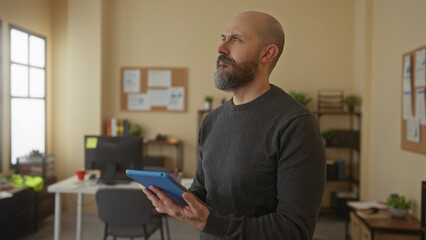 Fototapeta premium Bald man with beard holding tablet in office setting with shelves and desk in background conveying a professional workplace atmosphere.