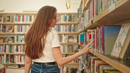 A confident young student walks through a university library, calmly browsing books on shelves. Woman in a modern library. Personal growth. Student life. Study habits. Personal development